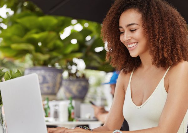 FAQ - A Young Woman Using Her Laptop While Smiling With a Plant Next to Her